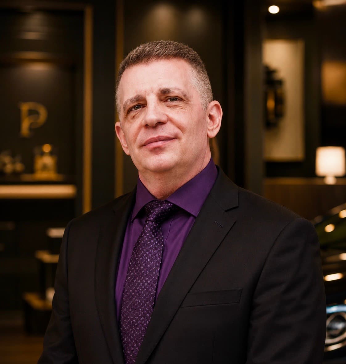 Middle-aged man in a black suit and purple shirt smiling in a sophisticated interior.