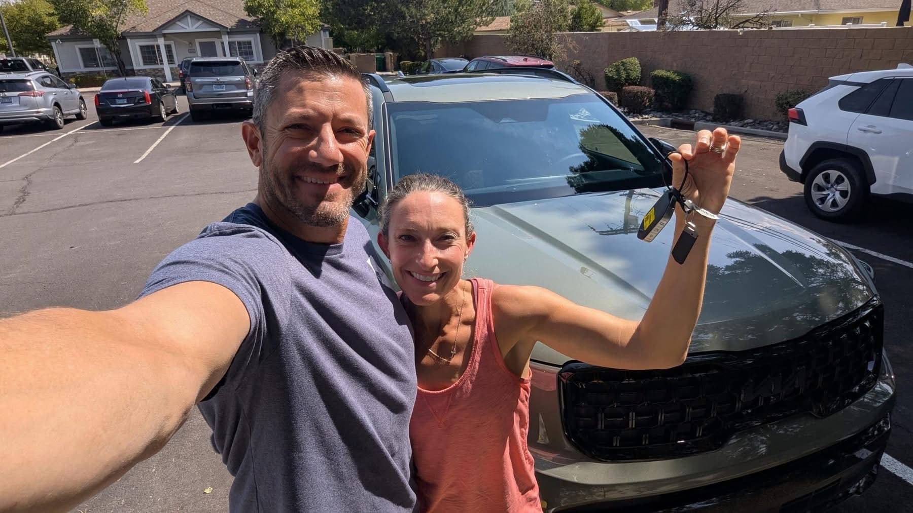 Smiling couple takes a selfie holding keys in front of their new olive green SUV.